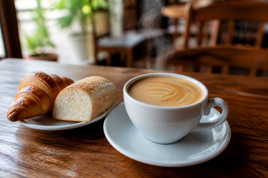 Uma xícara de café com leite fumegante sobre uma mesa de madeira em um café aconchegante, com pão ao lado e ambiente interno desfocado ao fundo.